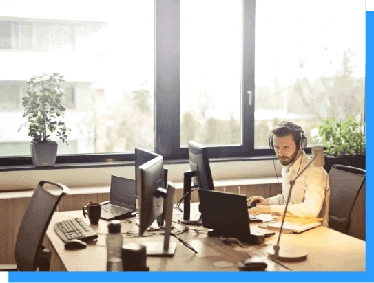 Modern office workspace with a man working at multiple computer stations, bright natural light from large windows, and greenery for a productive and comfortable work environment.