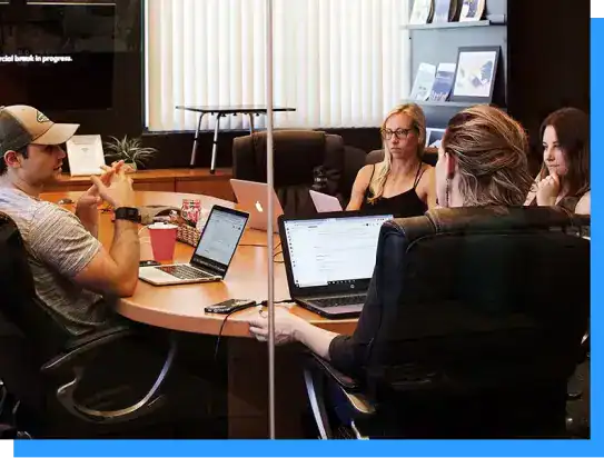 Modern office meeting with diverse professionals collaborating around a conference table with laptops and digital devices in a professional workspace.